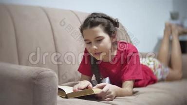 十几岁的女孩躺在沙发上看书。 躺在室内沙发上看书的女孩